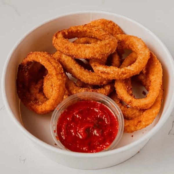 Golden-brown onion rings in a white bowl with a side of red dipping sauce.