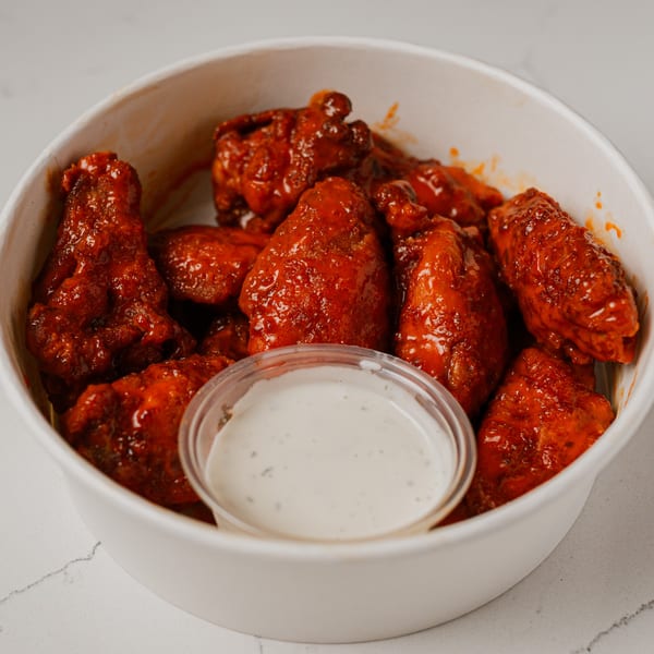 A white bowl filled with saucy red chicken wings and a small cup of ranch dressing.