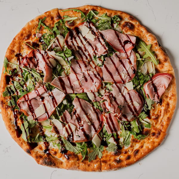 Pazza Pizza's prosciutto and arugula pizza, drizzled with balsamic glaze, on a white marble surface.